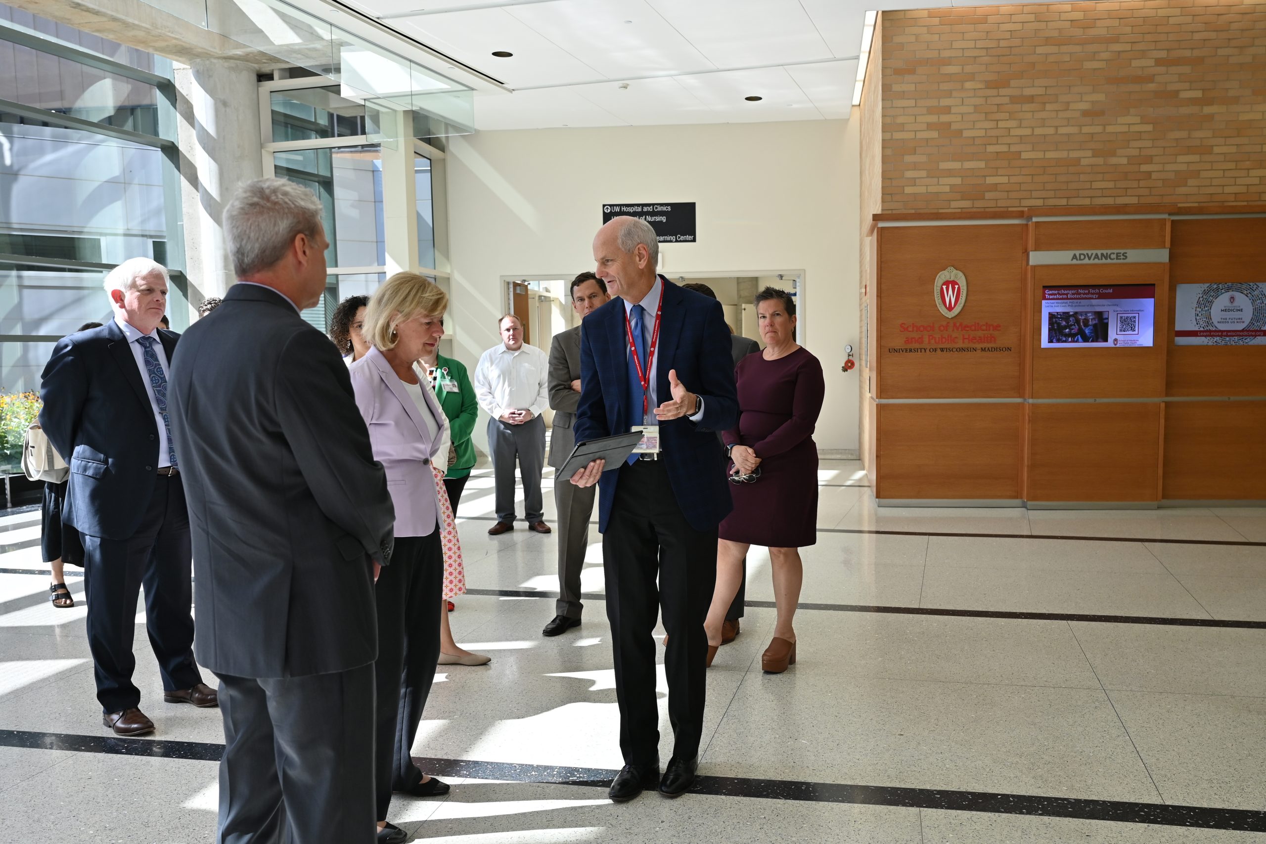 A group of people stand in a sun-lit lobby. At the forefront, Senator Baldwin listens as Dr. Grist speaks and points to an item on an iPad he holds. Several other people stand around them, listening. 