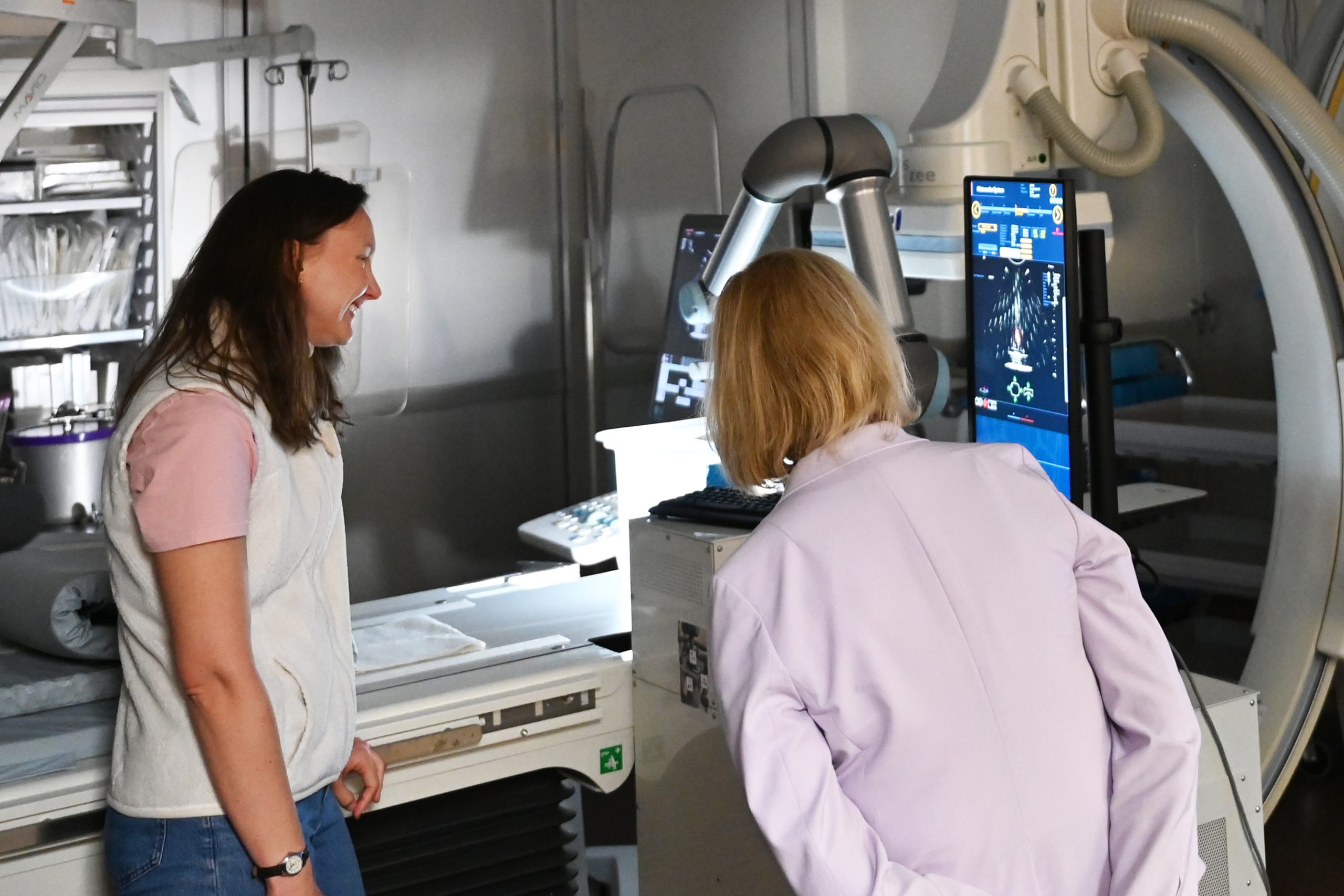 Meridith Kisting stand to the right speaking to Tammy Baldwin who faces away from the camera listening and observing the equipment Kisting is describing. In the background, is medical equipment including a large screen and scanning equipment.