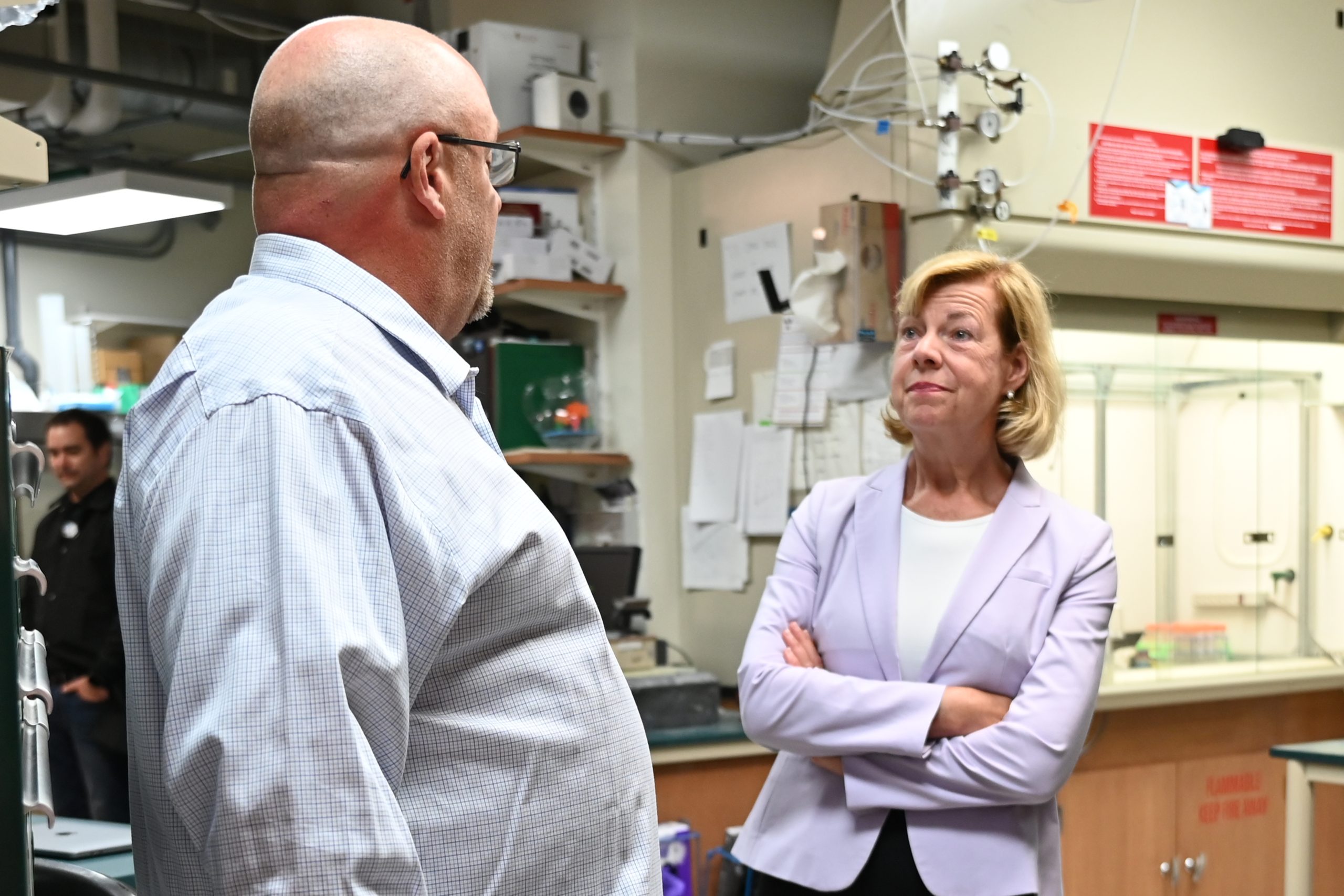 Senator Baldwin stand to the right facing Dr. Barnhardt as he speaks. In the background is lab equipment and personnel.