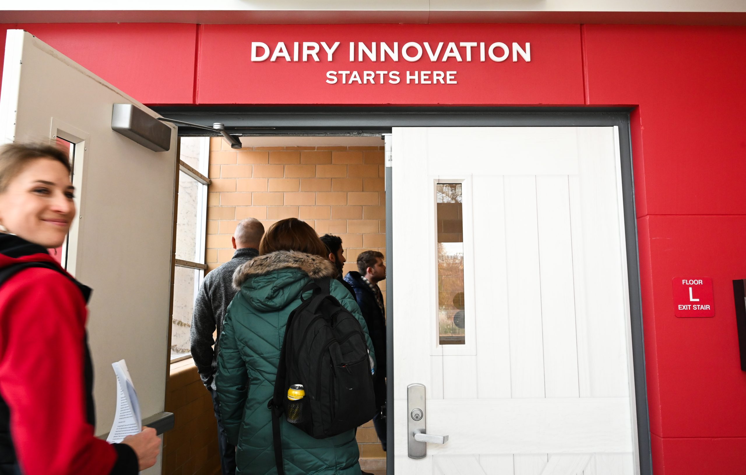 A group of people enters a doorway with a bright red frame and white door. Above the entrance, bold white text reads “DAIRY INNOVATION STARTS HERE.”