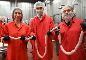 Three people wearing red lab coats, black gloves, and hairnets stand in a food processing facility holding a long, linked strand of raw sausage. The background shows stainless steel walls and industrial equipment.