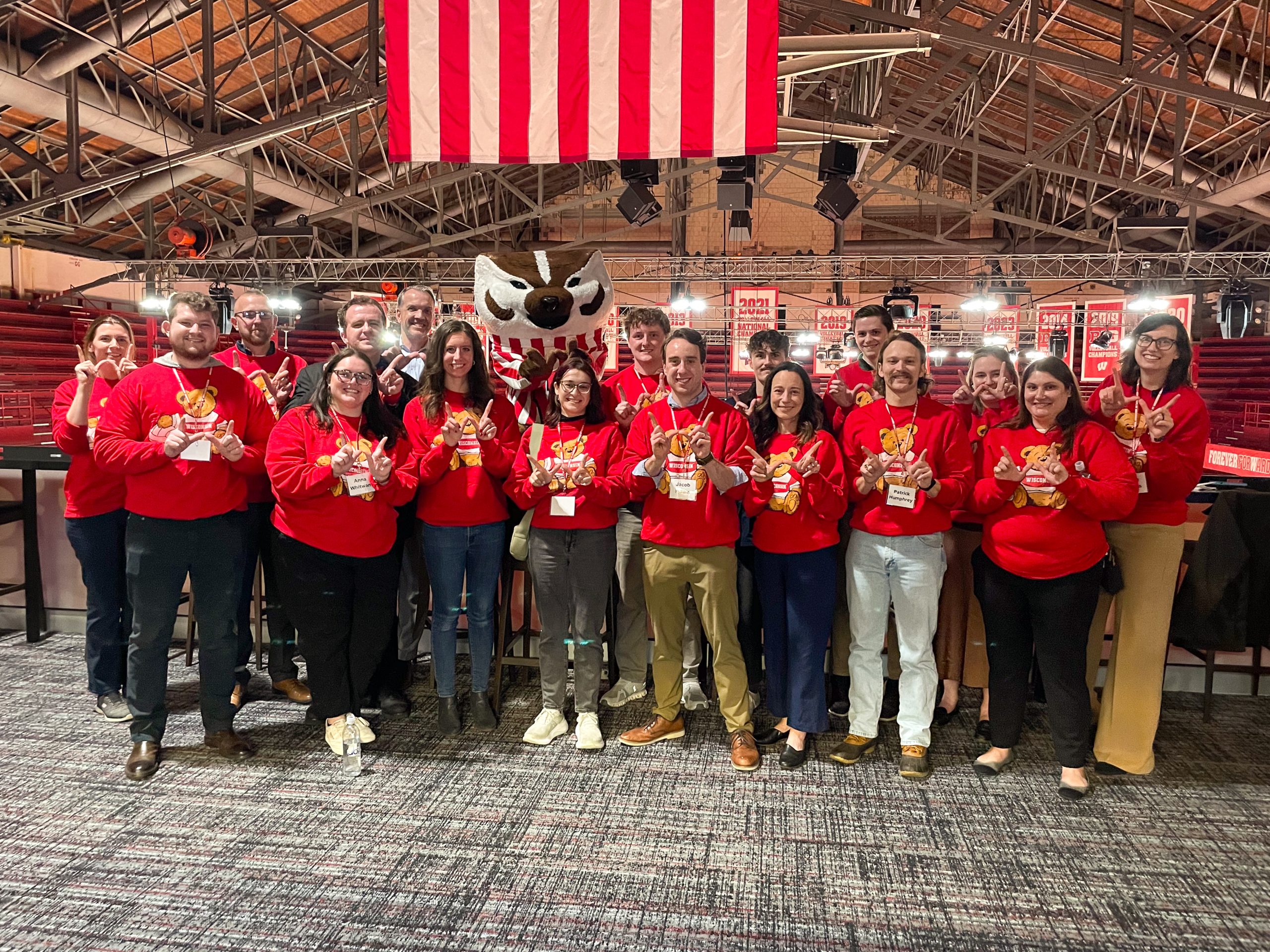 A group of people wearing matching red sweatshirts with a Bucky Badger design stand together inside the UW-Madison Field House, making a "W" with their hands.
