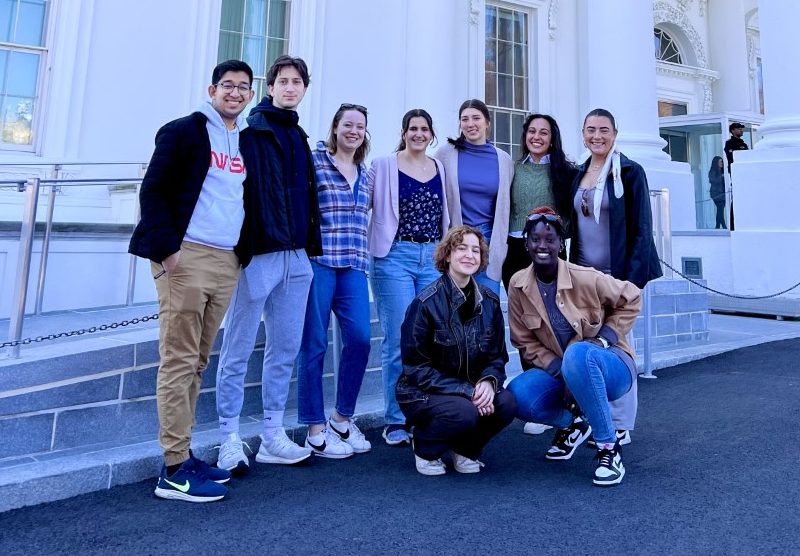 Nine young adults pose for a group photo outdoors in front of a white building with large neoclassical columns.