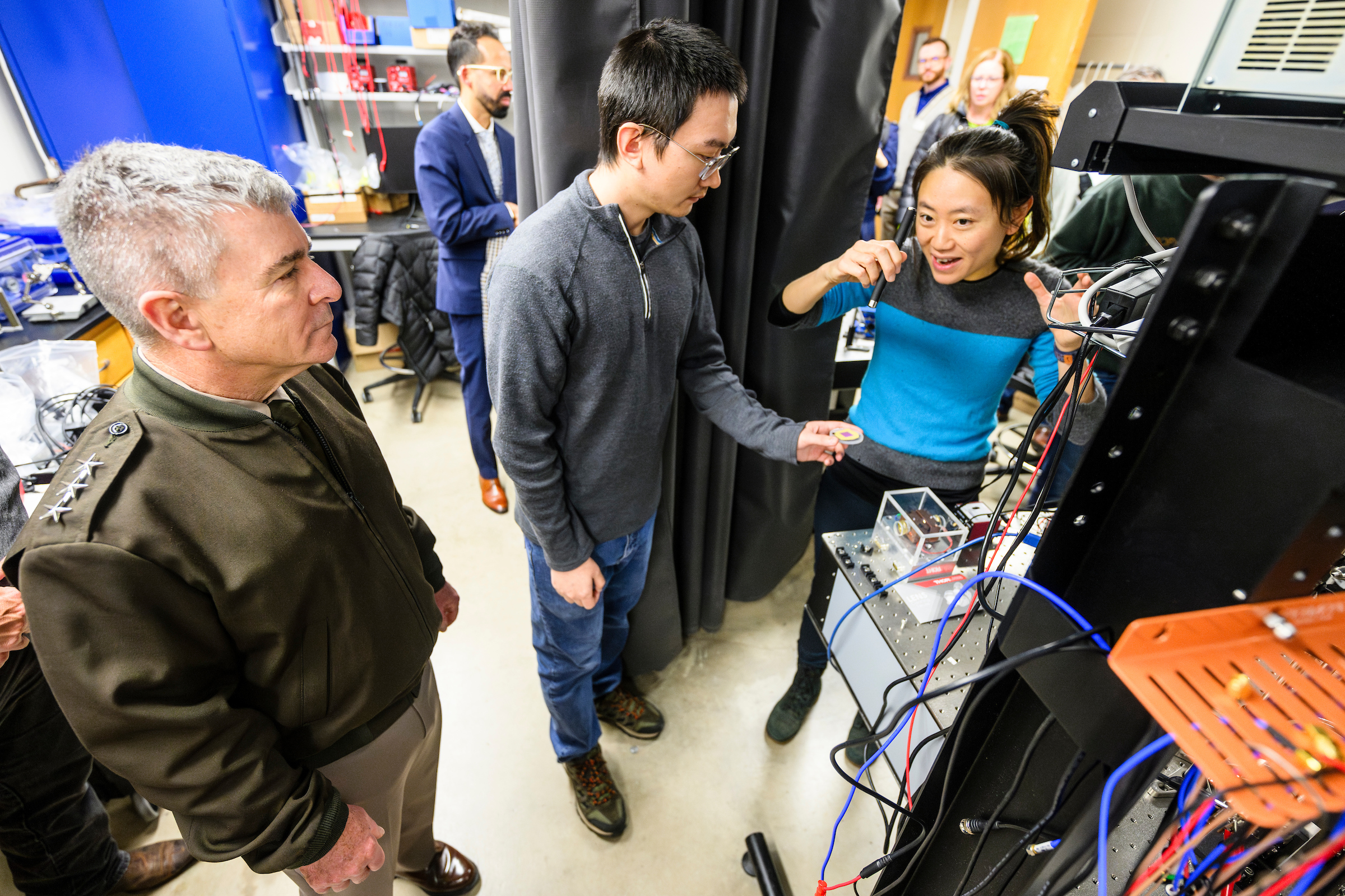 Three individuals examine equipment in a laboratory setting, with one person explaining the functioning of the device to the others.
