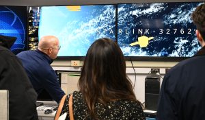 Several people are gathered in a control room, looking at large monitors displaying a satellite image of Earth with the text “STARLINK-32762” and a yellow satellite graphic. The room has multiple screens, computer equipment, and bright overhead lighting.