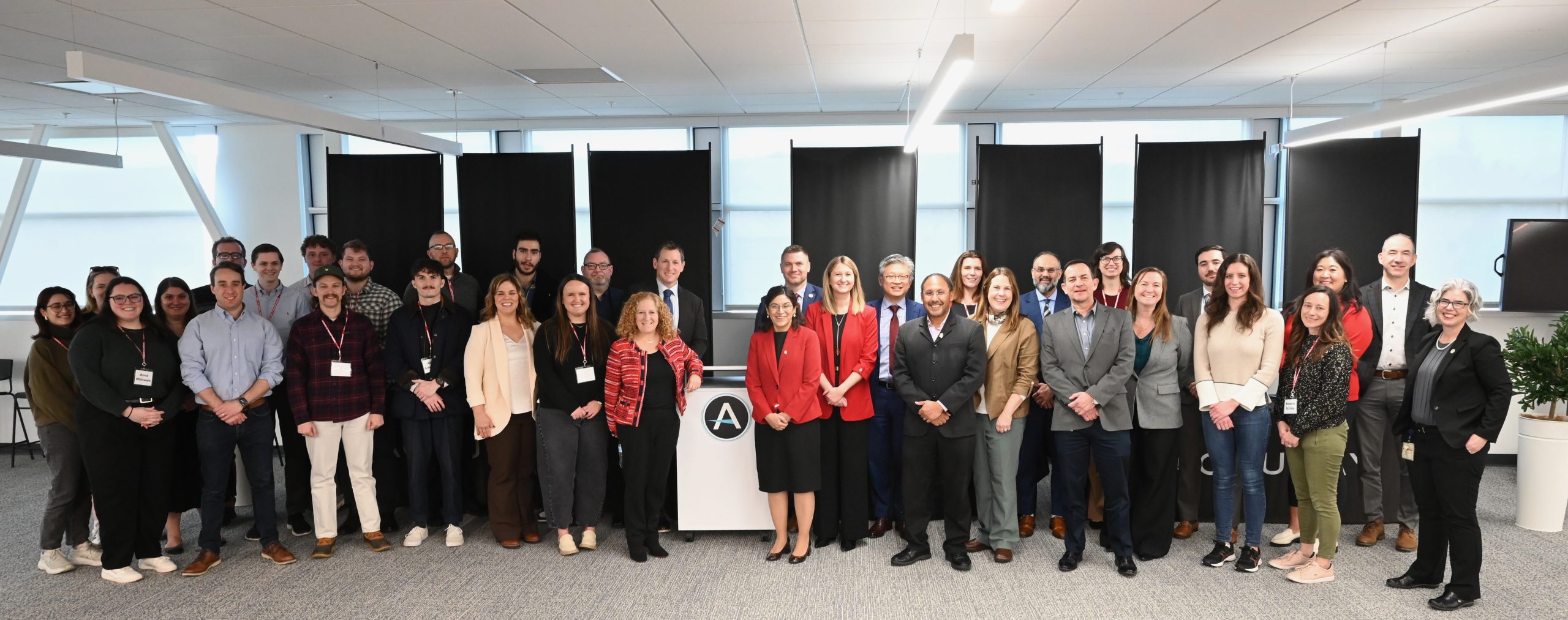 A large group of people stand together in a modern office setting with bright overhead lighting and gray carpeted floors. They are arranged in two rows behind a white podium featuring a circular logo with the letter “A.” Several individuals wear business attire, while others are dressed in business casual clothing. Behind the group are tall windows covered with black shades, and a large white planter with green foliage is visible on the right side of the image.